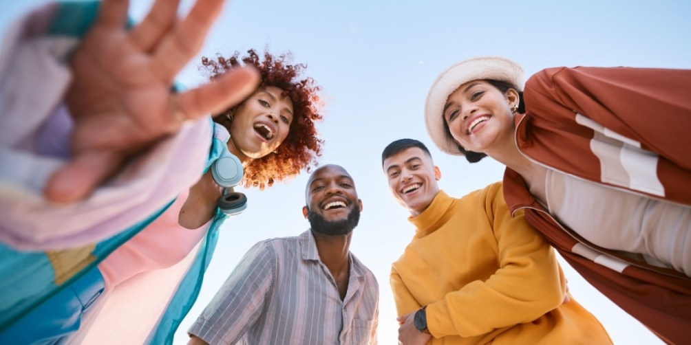 Four smiling friends looking down at the camera against a clear blue sky, enjoying spring break safely while spending time outdoors as a group.