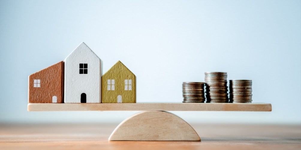 Three miniature wooden houses and stacks of coins balanced on a wooden seesaw, visually illustrating how insurance rates are determined based on risk assessment.