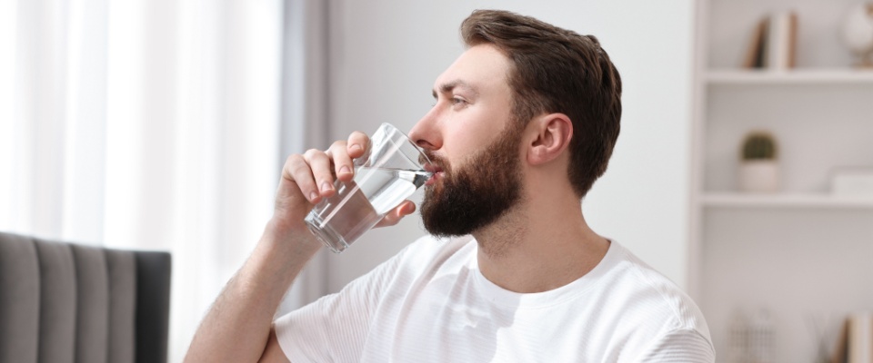 Man in a white t-shirt sitting at a dining table and drinking a glass of water in a bright, modern home, staying hydrated to maintain good health as a summer tip.