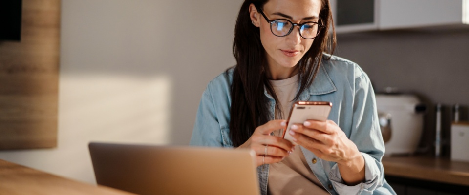 Woman using her smartphone at a kitchen table to create and store an emergency contact list for quick access during urgent situations.