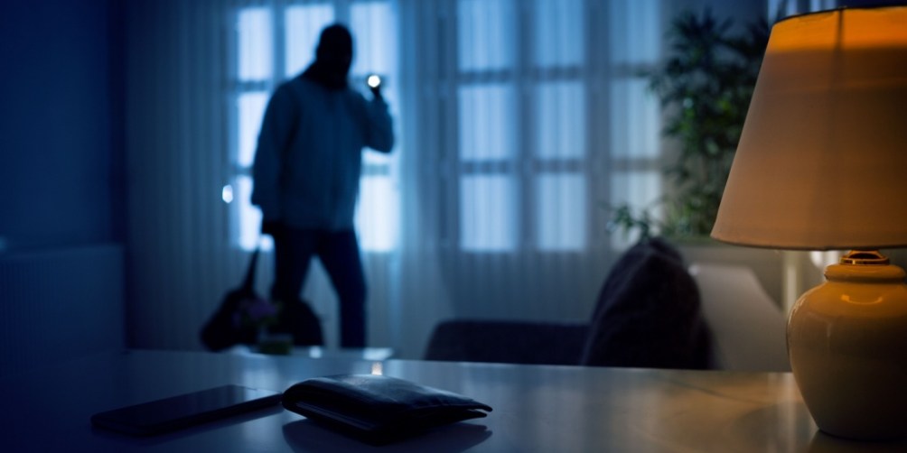 A burglar sneaking into a dimly lit apartment through the window at night, with a wallet and smartphone visible in the foreground, highlighting the importance of learning how to deter burglars and protect your valuables.