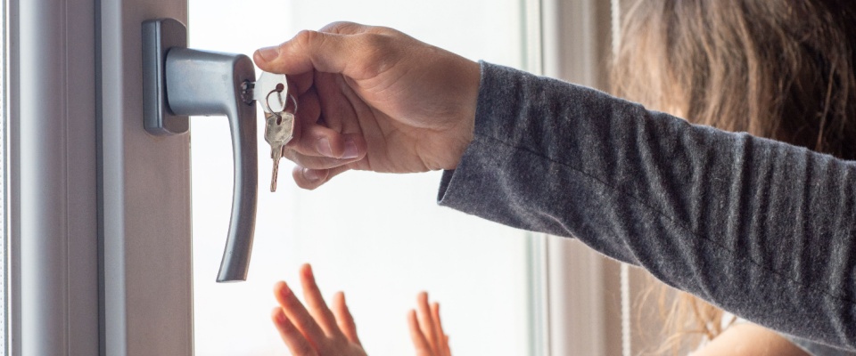 Person locking a window with a key while a child stands nearby, illustrating practical steps on how to deter burglars from breaking in.
