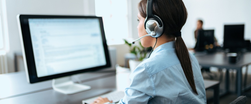 A renters insurance agent wearing a headset assists a client on a computer in a modern office, assisting a client in figuring out how much renters insurance they need.