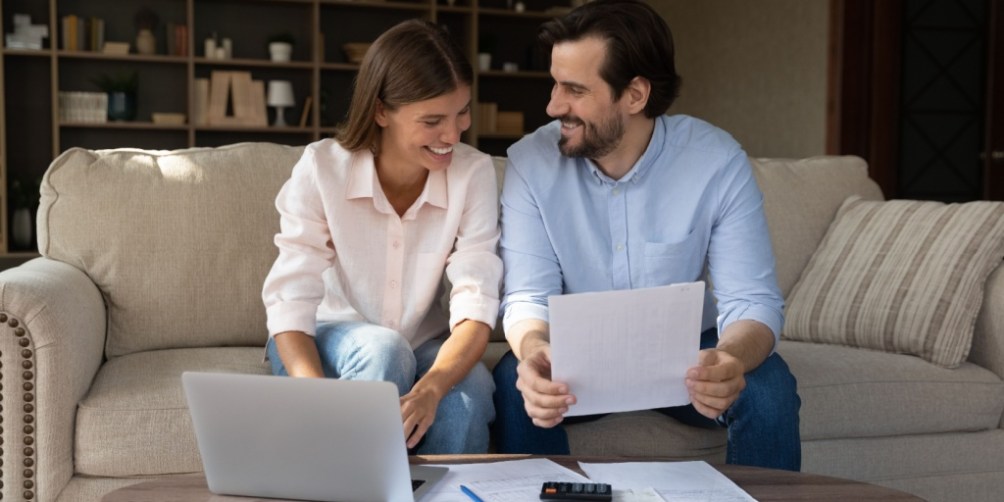 A couple sits together on a beige sofa, reviewing documents and using a laptop with bills and a calculator on the coffee table, thoughtfully discussing how much renters insurance they need.