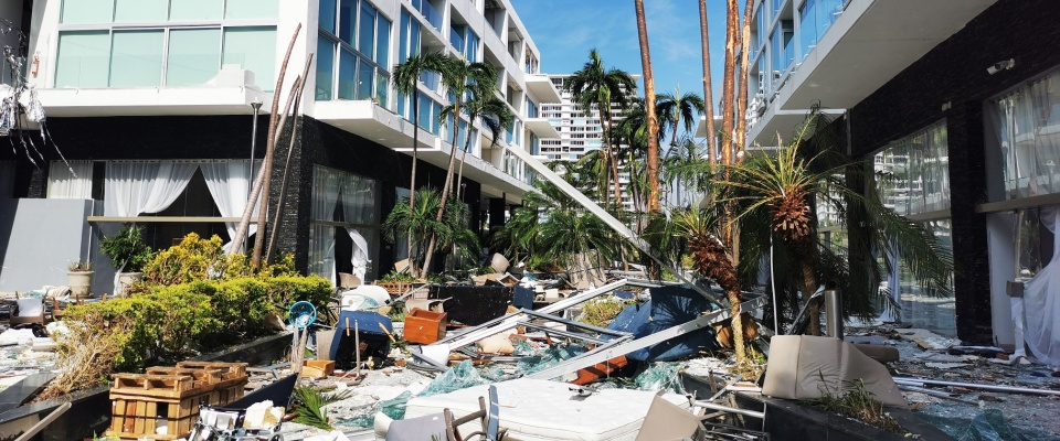 Debris and broken palm trees litter the ground between modern apartment buildings after a destructive storm, illustrating potential types of damage to be covered by renters insurance in the aftermath of a hurricane. 