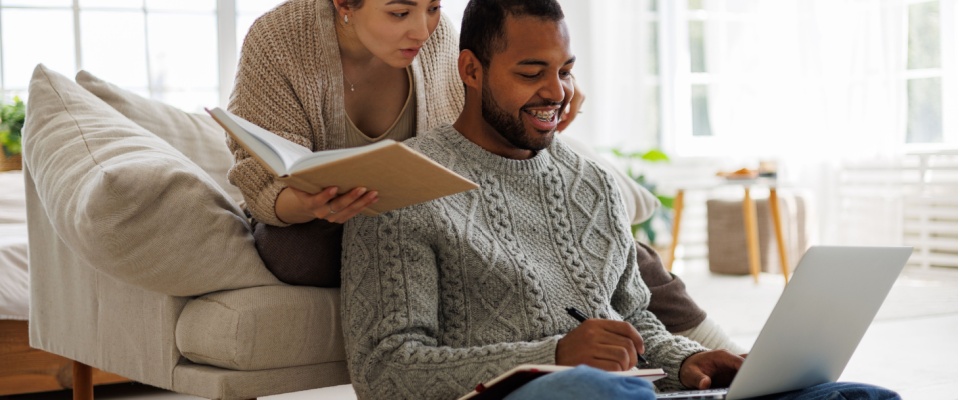 A man in a grey cable-knit sweater works on a laptop with notes in hand while a woman stands nearby holding a notebook in a bright, modern living room, reviewing their renters insurance coverage for hurricane damage.