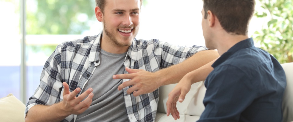 Two young men sit on a couch engaged in a thoughtful conversation, considering how important communication is when choosing roommates to move in with.