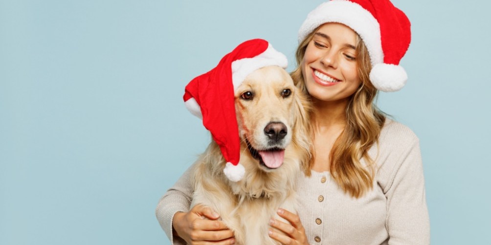 Smiling woman posing with her dog in matching Santa hats, celebrating joyful and pet-friendly holidays together.
