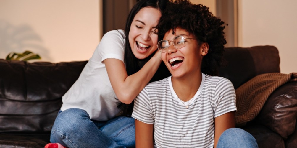 Two young women sit together on a dark leather sofa in a cozy living room, illustrating the importance of sharing and discussing questions to ask potential roommates before moving in together.