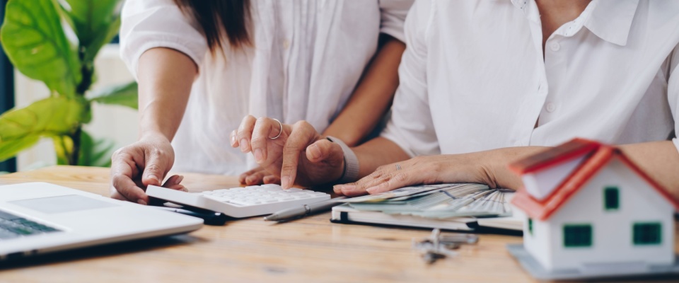 Two roommates in white shirts review financial documents and use a calculator at a table with a miniature house and laptop nearby, discussing important questions about splitting utilities.