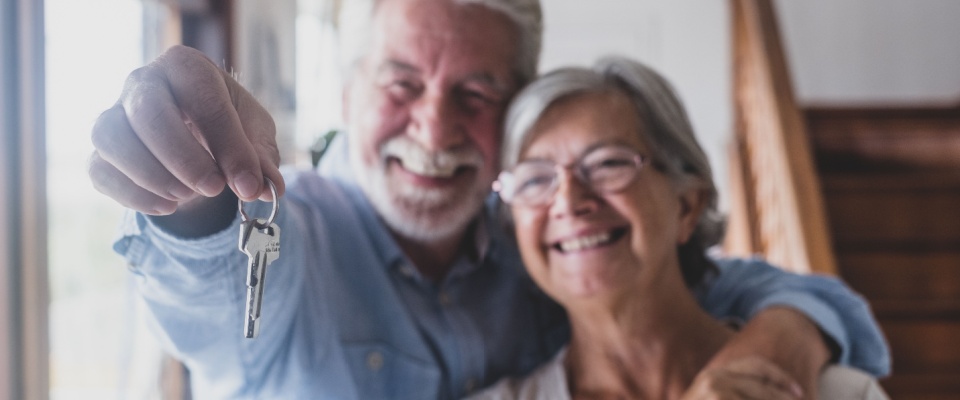 An older couple stands together at home, holding up a house key in front of a staircase, visually celebrating the transition to a more manageable living space and highlighting the positive aspects of downsizing for seniors.