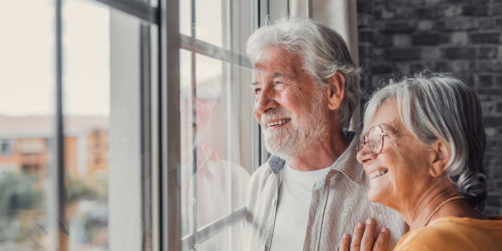 An senior couple stands together by a window in a cozy apartment, quietly looking outside and reflecting on the implications of downsizing.