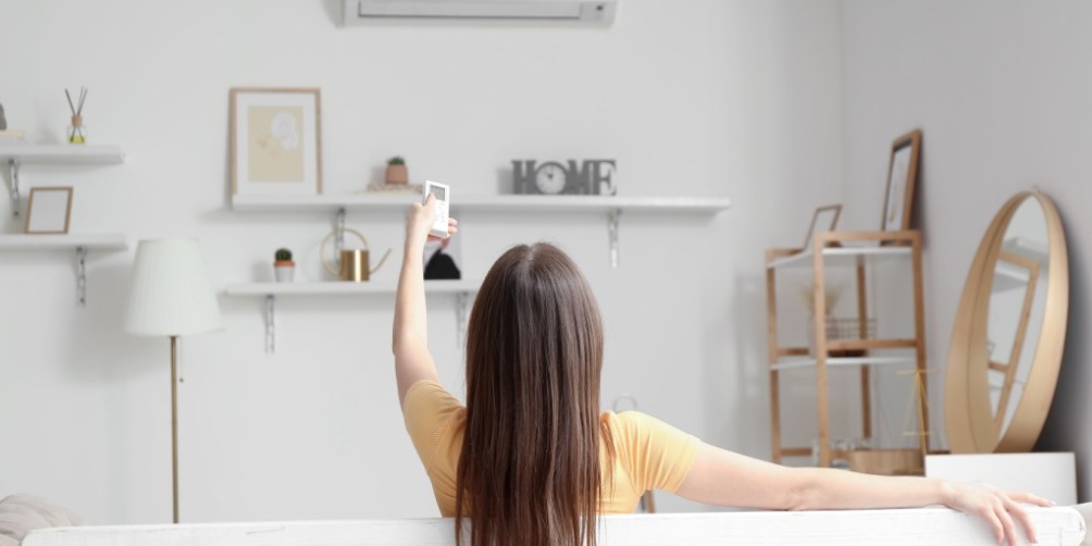 Woman sitting on a sofa in a bright living room, using a remote to adjust the air conditioner, illustrating how to lower the electric bill in summer by optimizing thermostat settings.