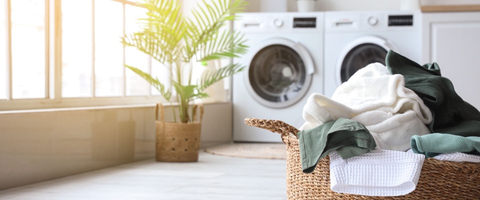 Bright laundry room with sunlight streaming through large windows, featuring a basket of clothes in front of energy-efficient washing and drying machines, to be used off-peak hours to lower the electric bill in summer.