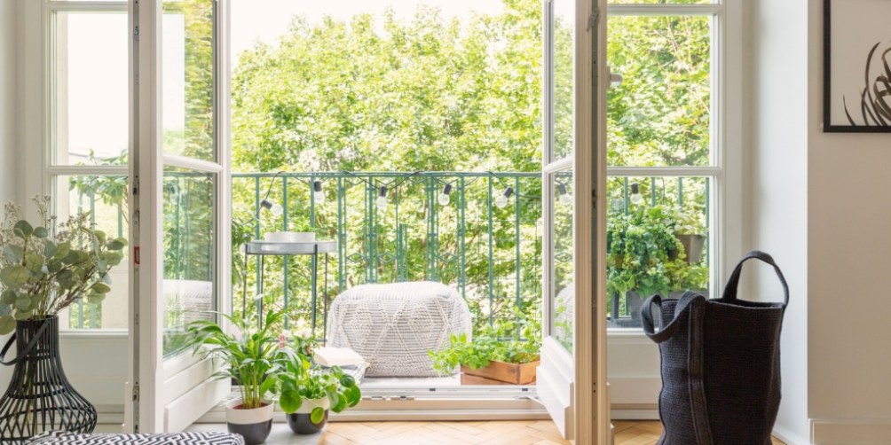 Bright apartment living room with open French doors leading to a balcony filled with lush green plants, showcasing how to prepare an apartment for summer.