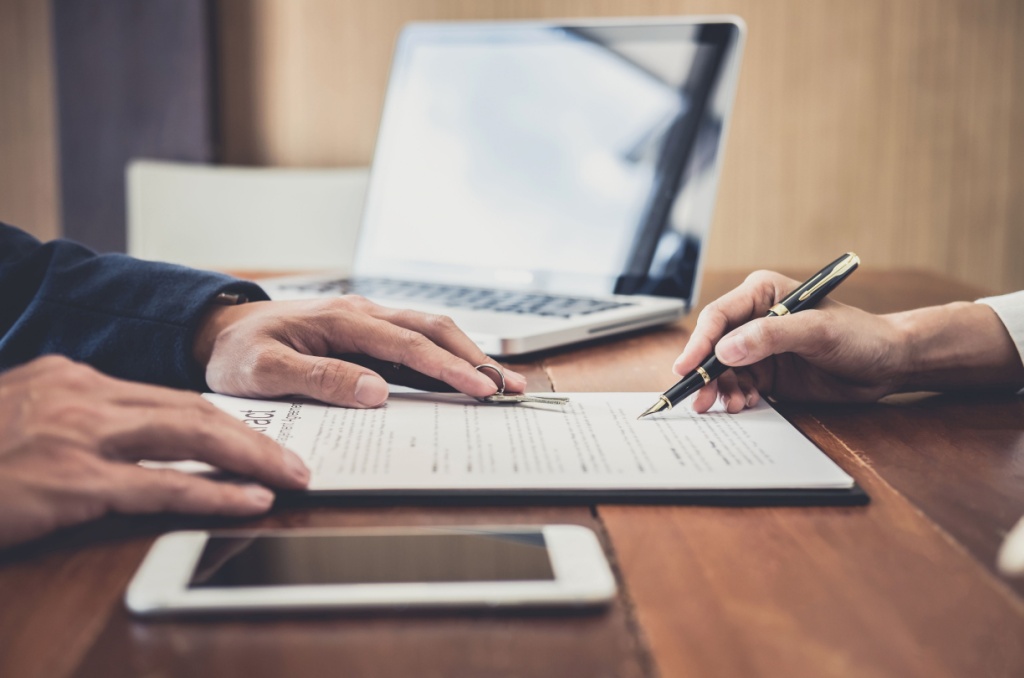 Two people signing a lease agreement at a wooden table with a laptop and tablet nearby, after reviewing important questions to ask before signing a lease.