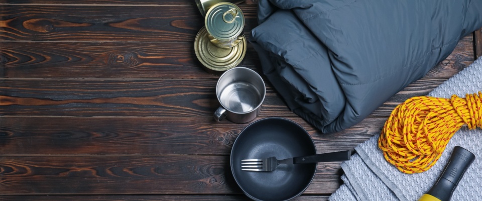Camping sleeping bag, metal cookware, water bottle, insulated mug, rope, and towel neatly arranged on a wooden surface, illustrating important non-electrical essentials to include in a power outage emergency kit.