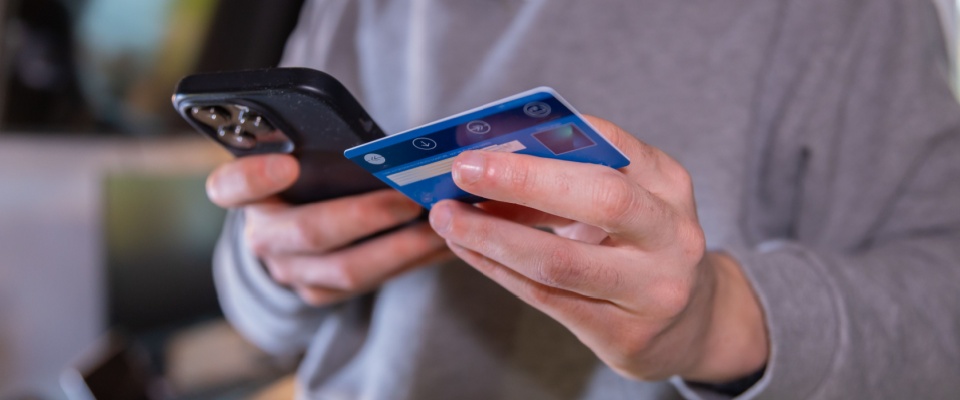 Person in a gray sweatshirt holding a smartphone and a blue credit card, demonstrating the importance of being cautious with online payments and personal information as part of learning how to avoid rental scams.