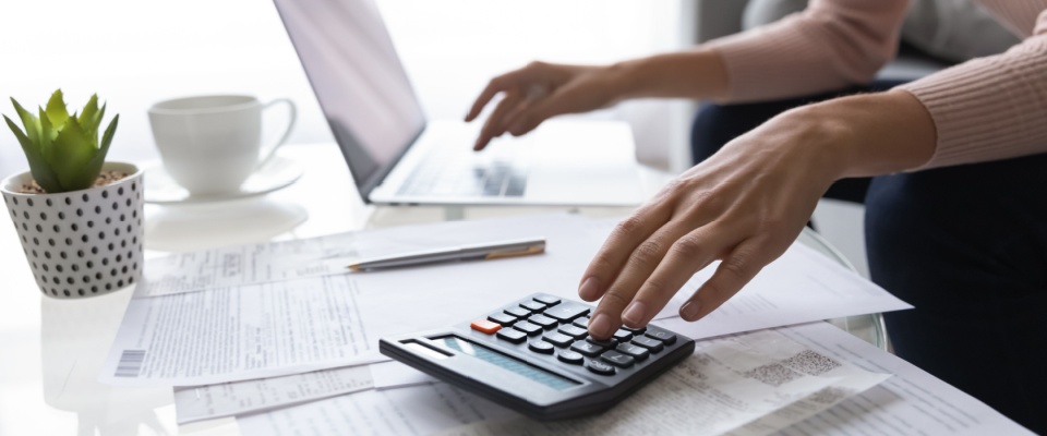 Person in front of a white coffee table, with a laptop, calculator, apartment lease agreement, a potted plant and a white cup, calculating rent costs. 