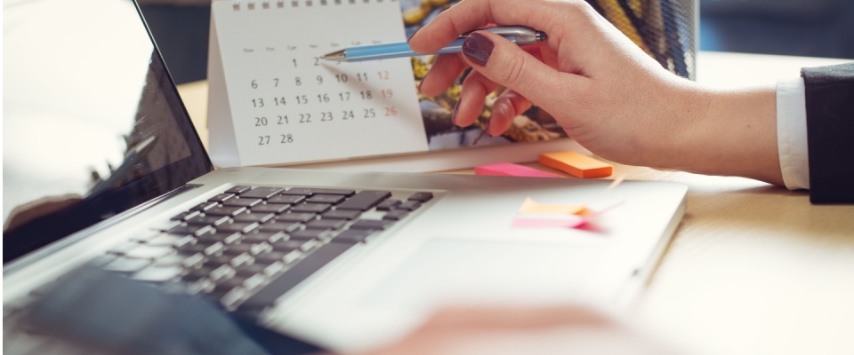 Hand holding a pen while pointing at a calendar next to a laptop and colorful sticky notes, picking a date for the renters insurance cancellation.
