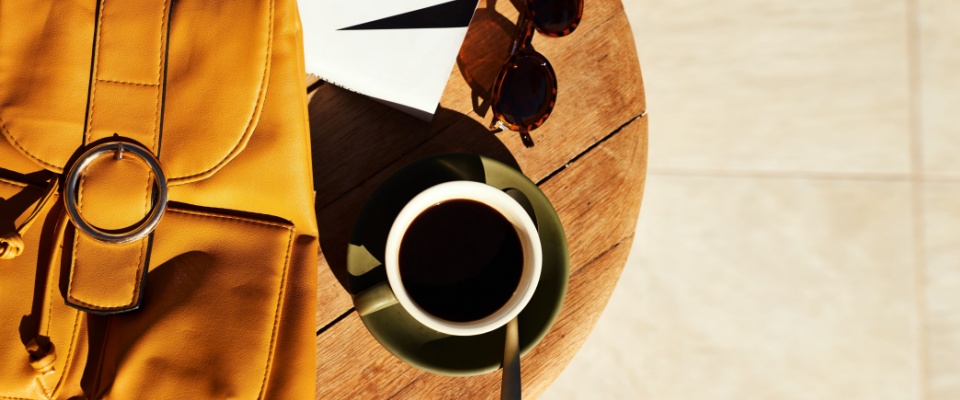Yellow backpack, cup of coffee, sunglasses, and book arranged on a wooden table in sunlight, representing a lesser-known renters insurance benefit of off-premises coverage for personal belongings outside the renter’s apartment.