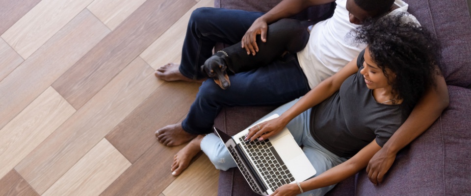 A couple relaxing together on a purple couch with their small black dog, while using a laptop to search for pet-friendly rents.