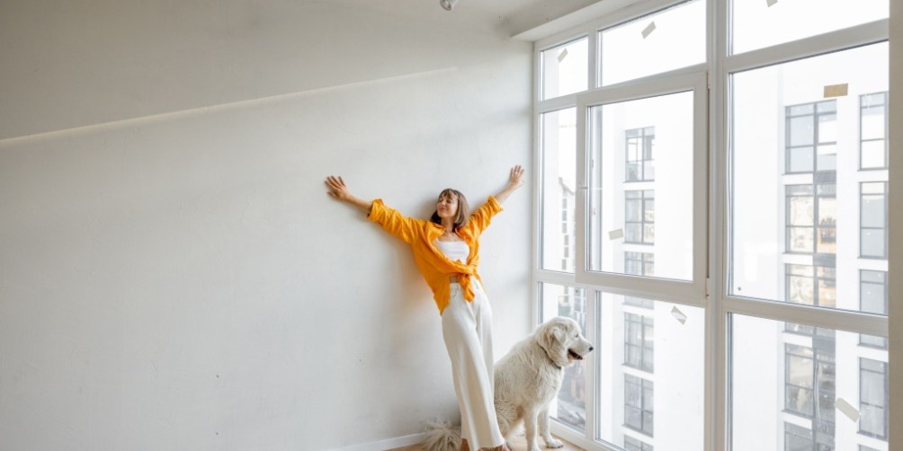 Bright modern apartment interior with white walls, featuring a person standing near a large window alongside a large white dog, feeling joyful after successfully renting with pets.