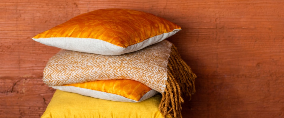 Stack of cozy orange and yellow pillows with a soft herringbone throw blanket, set against a rustic terracotta wall, showcasing a simple decor idea for fall.