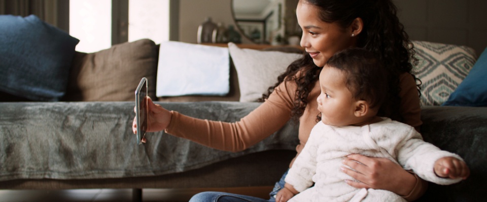 Mother sitting on the floor holding her baby and taking a selfie on a smartphone, representing family awareness and digital privacy while staying safe online.