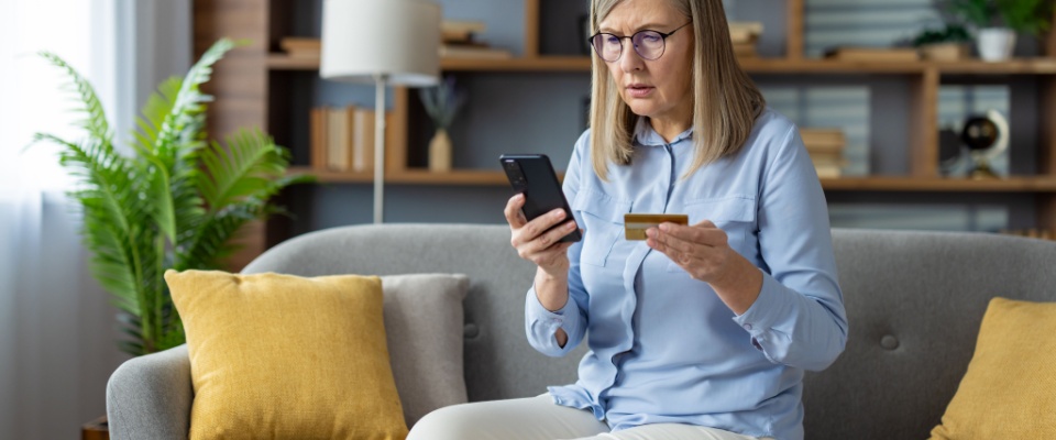 Older woman sitting on a couch holding a smartphone and credit card, being concerned about online security and phishing risks.