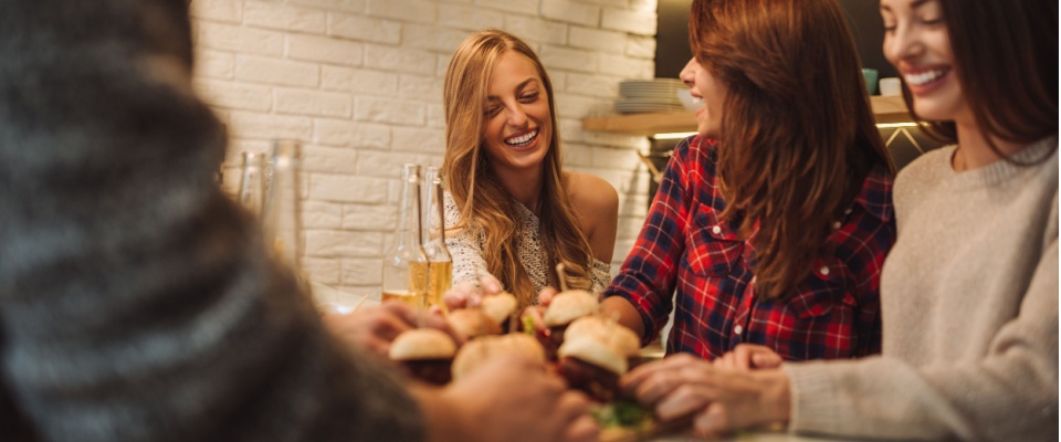 Group of friends in gathered in an apartment around a table, enjoying burgers and drinks, while following safe practices during the 4th of July.