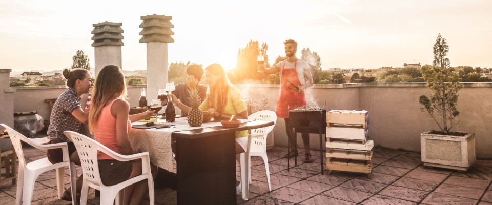 Group of friends enjoying an outdoor meal on a rooftop patio at sunset, with one person grilling near the table, while following 4th of July grilling safety tips.