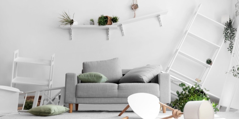 A living room with a gray sofa, toppled shelves, scattered plants, and an overturned lamp visually demonstrates earthquake damage, raising the question of whether renters insurance covers earthquakes for protecting belongings.