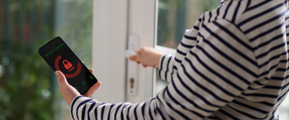 Person in a striped shirt using a smartphone with a security app and locking a patio door, illustrating how to protect your home while on vacation.