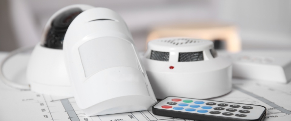 Collection of home security safety gadgets including a smoke detector, motion sensor, and remote control placed on a table.