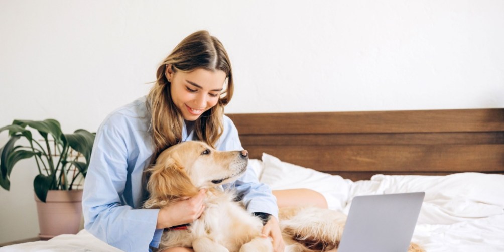 Woman sitting on a bed in a bright bedroom, gently hugging her golden retriever next to an open laptop, illustrating a common scenario for pet owners wondering if renters insurance covers pet damage.