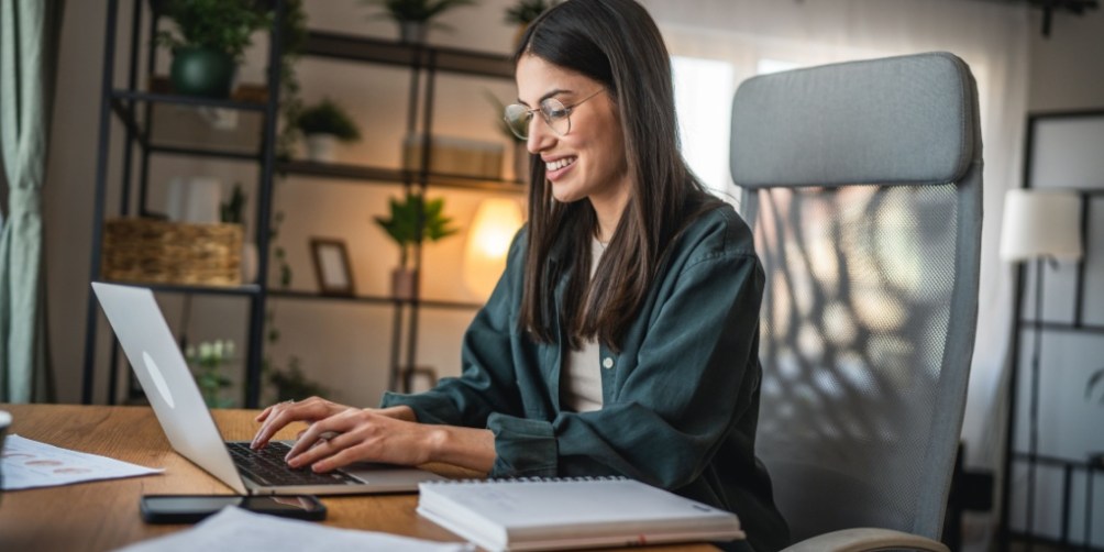 Woman working on a laptop at a desk in a bright, organized home office, considering work-from-home safety by using an ergonomic setup.
