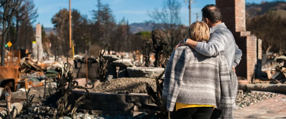 Couple embracing while viewing the aftermath of a wildfire, surrounded by burnt homes and scorched landscape, illustrating the importance of renters insurance wildfire coverage for recovery after fire damage.