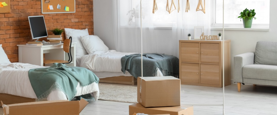 Bright college dorm room with two beds, a study desk, moving boxes, and a wardrobe, illustrating how student renters insurance works for on-campus versus off-campus living.
