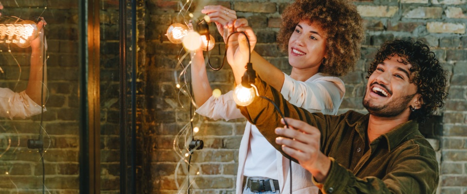 Two people hanging decorative string lights indoors while hosting a New Year’s Eve party, preparing festive and safe home decorations.