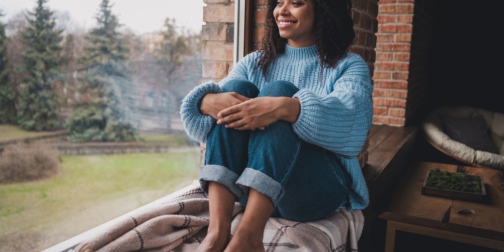 Smiling woman sitting by a well-insulated window wrapped in a blanket, enjoying warmth after taking steps to winterize her apartment.