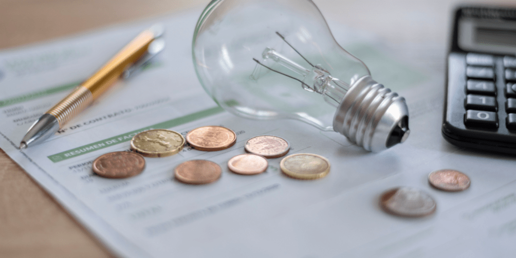 Electricity bills covered by several coins, a light bulb, calculator, and pen on a desk.