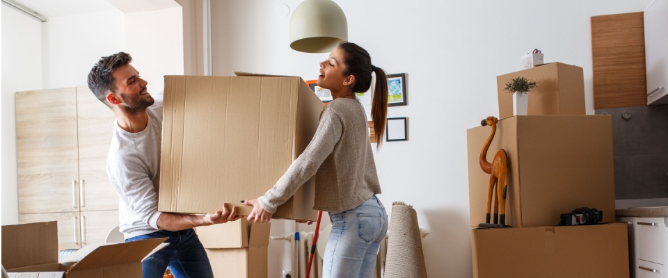 A man and woman carrying a large cardboard box into a room filled with other moving boxes, suggesting the excitement of renting an apartment at the best time.