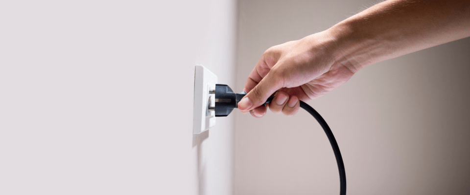 Close-up of a hand inserting a black electrical cord into an outlet on a white wall, highlighting the importance of handling plugs properly as an electrical safety tip.