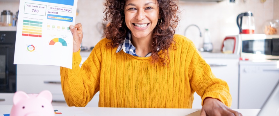 Woman in a bright kitchen holding a printed sheet with high credit score and financial information, wearing a yellow sweater, with a pink piggy bank and paperwork on the table.