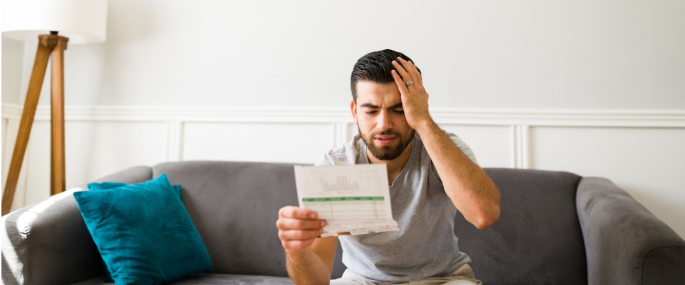Man sitting on a gray sofa with a teal pillow, looking at a bill in a bright, modern living room of an all-bills-paid apartment, worries about the electricity bill cost.