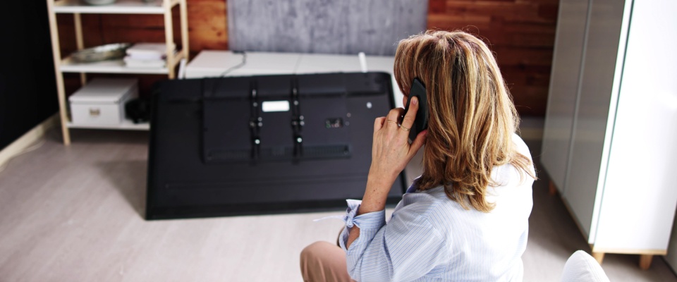 Woman on the phone sitting in a modern living room, looking at a large flat-screen TV that has fallen and broken, visually demonstrating a real-life scenario where understanding your renters insurance deductible is crucial for handling damage claims.