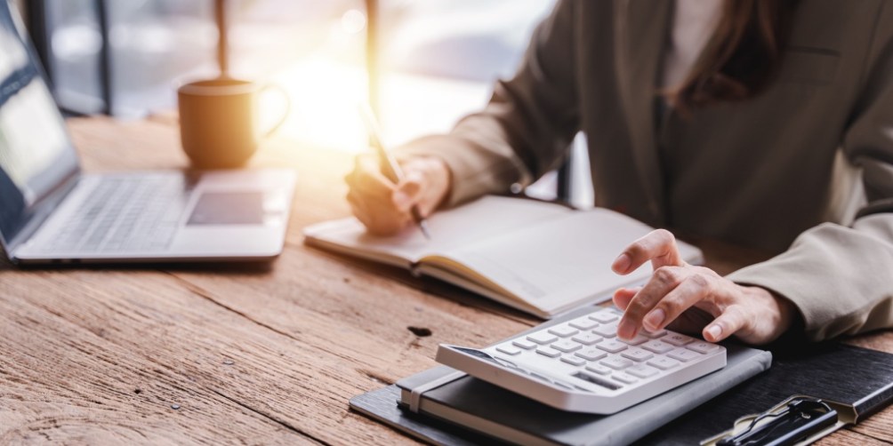 Person sitting at a wooden desk using a calculator and writing in a notebook, with a laptop and coffee cup nearby, calculating a renters insurance deductible.