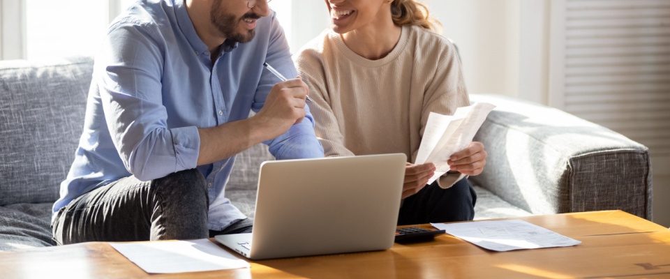Smiling couple sitting together at a coffee table, reviewing paperwork and using a laptop to review their renters insurance deductible.