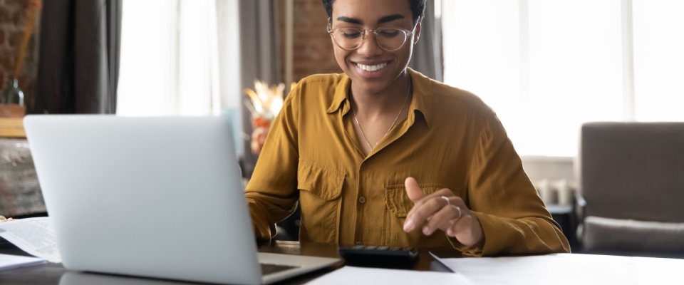 Smiling young woman working on a laptop and calculator at home, figuring out the low cost of renters insurance.